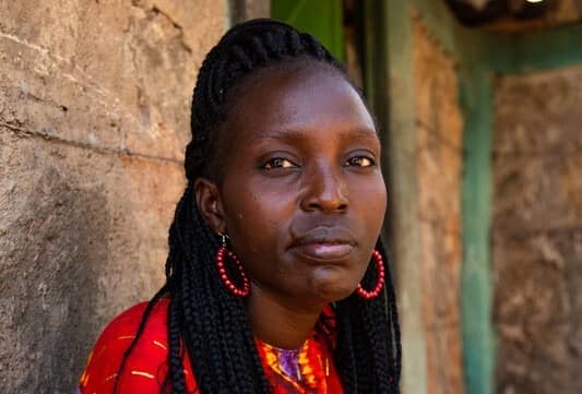 Portrait of a person with braided hair and red earrings, wearing a vibrant shirt, standing by a rustic wall.
