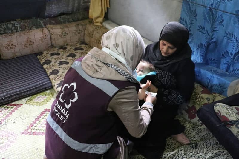 Healthcare worker assisting mother and baby in a modest living space.