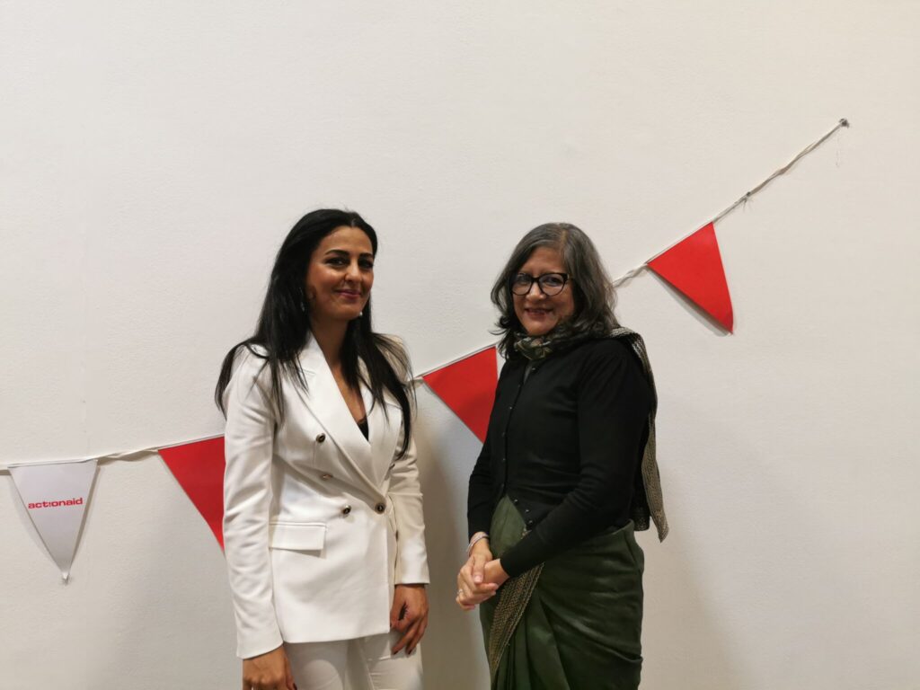 Two women smiling in front of ActionAid bunting on a white wall, one in a white suit, the other in a green sari.