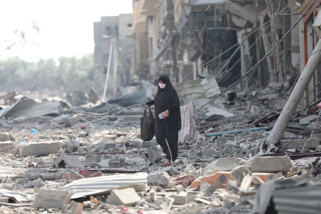 Woman walking through rubble in ruined street, wearing mask and carrying bags amidst destruction.