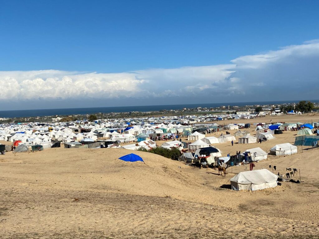 Tents in a sandy refugee camp under a blue sky with distant cityscape and sea horizon.