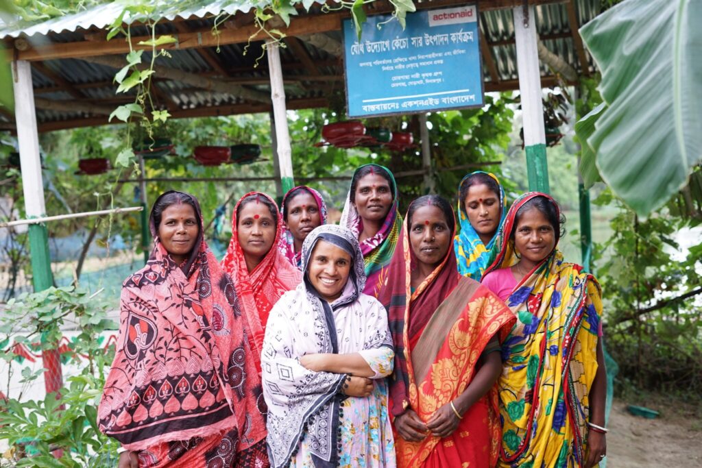 Group of women in colorful sarees smiling, standing outside a community center surrounded by greenery.
