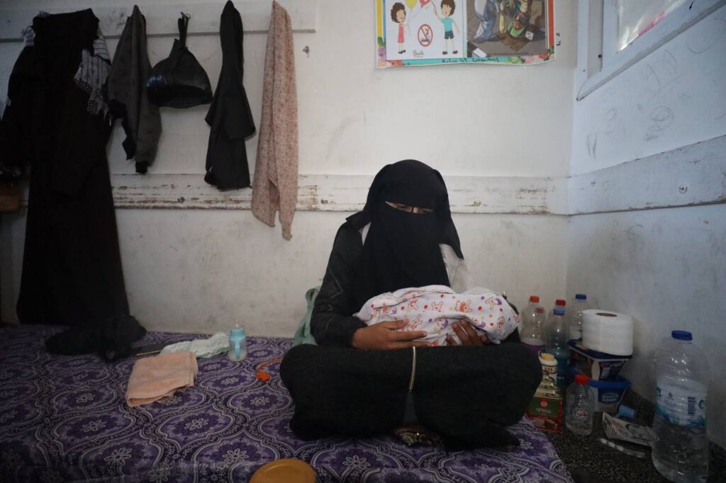 Woman in a niqab sits on a bed holding a baby, surrounded by household items, highlighting a domestic setting.