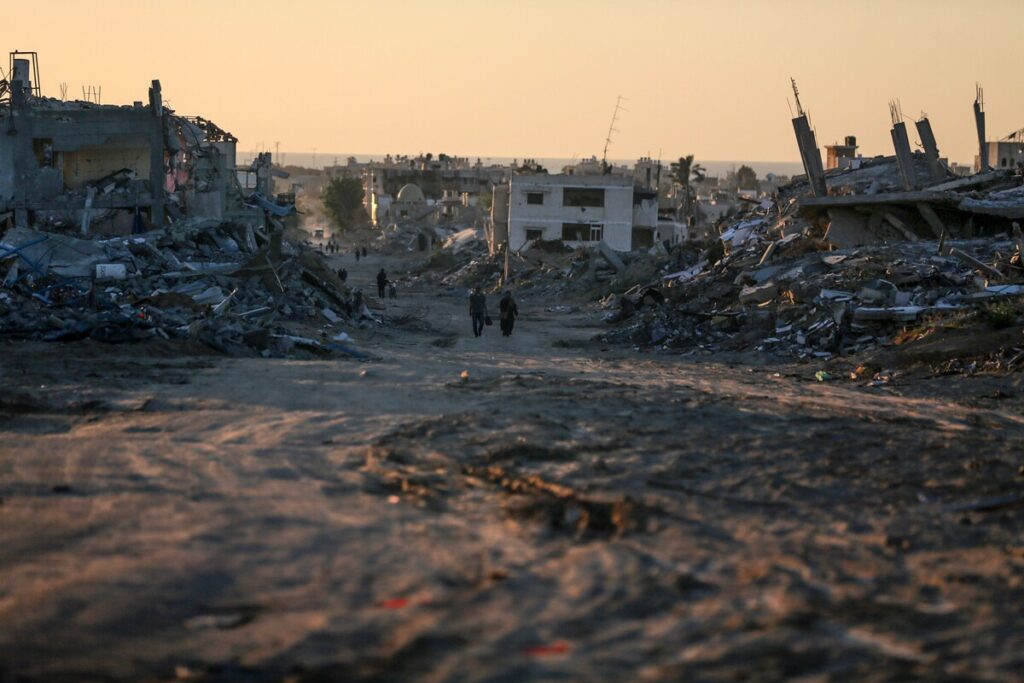 Rubble-strewn street at sunset, with distant figures walking amid war-torn buildings.
