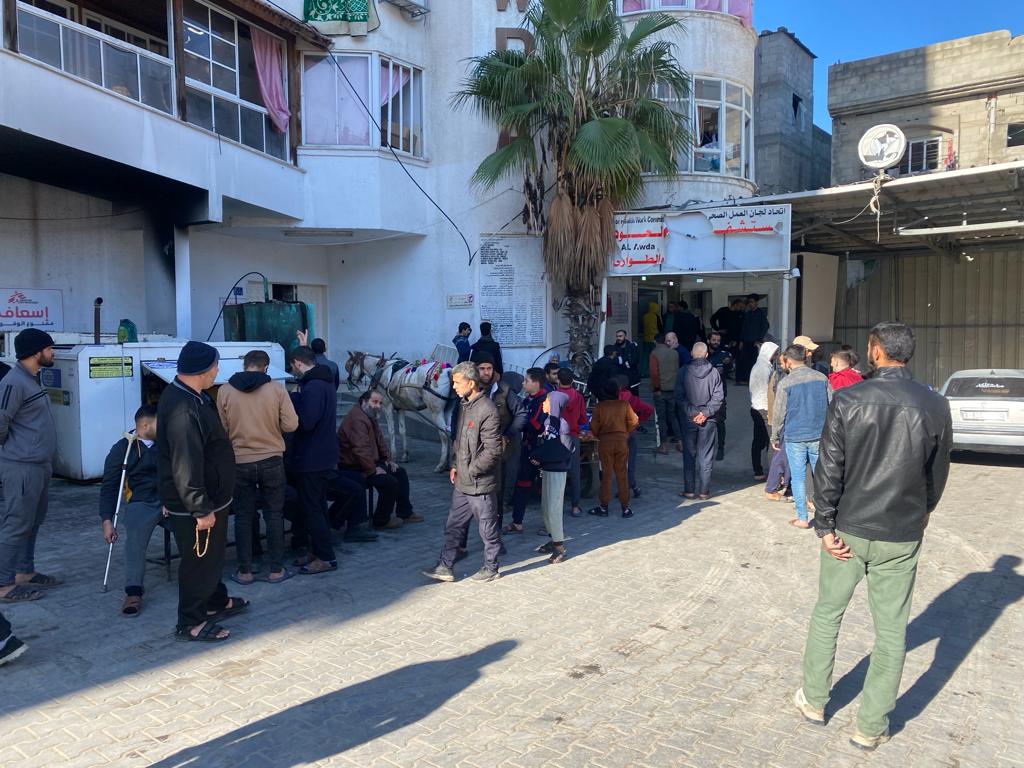 People waiting in line outside a building on a sunny day, with a palm tree and a horse-drawn cart nearby.