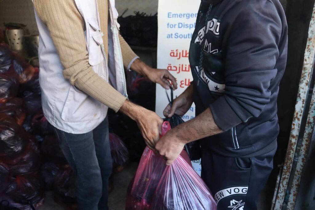 Two people handling a food package in a relief center for displaced persons, with bags stacked in the background.