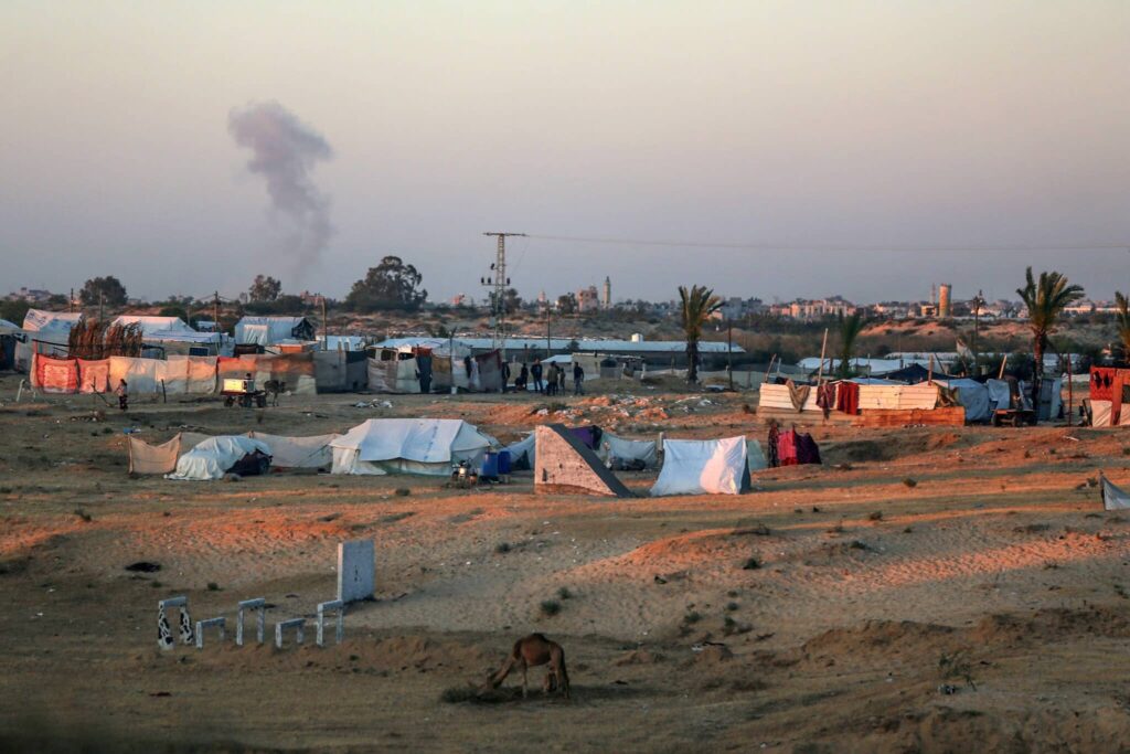 Temporary shelters in a refugee camp with distant smoke cloud under an evening sky.