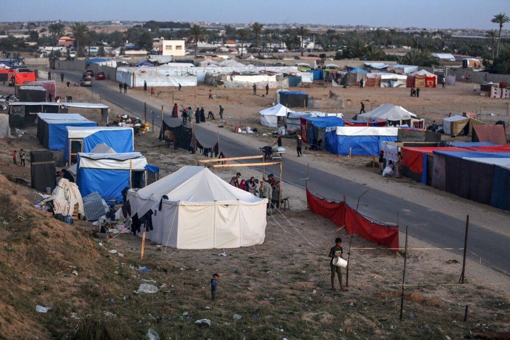 Temporary shelters in a refugee camp with families, tents, and surrounding landscape, under a clear sky.