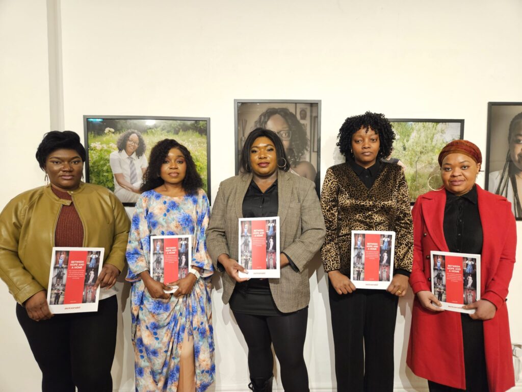 Group of five women holding books at an event, standing against a backdrop of framed portraits.