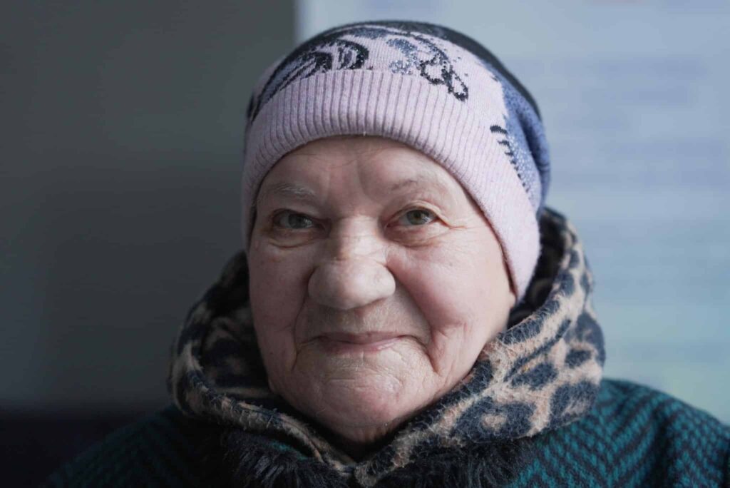 Elderly woman in a warm hat and coat, smiling gently against a neutral background.