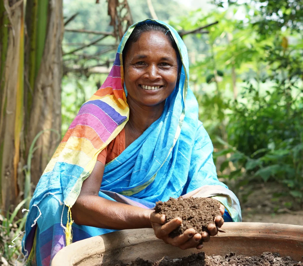 Smiling woman in colorful sari holding soil, demonstrating sustainable organic farming in a lush garden setting.