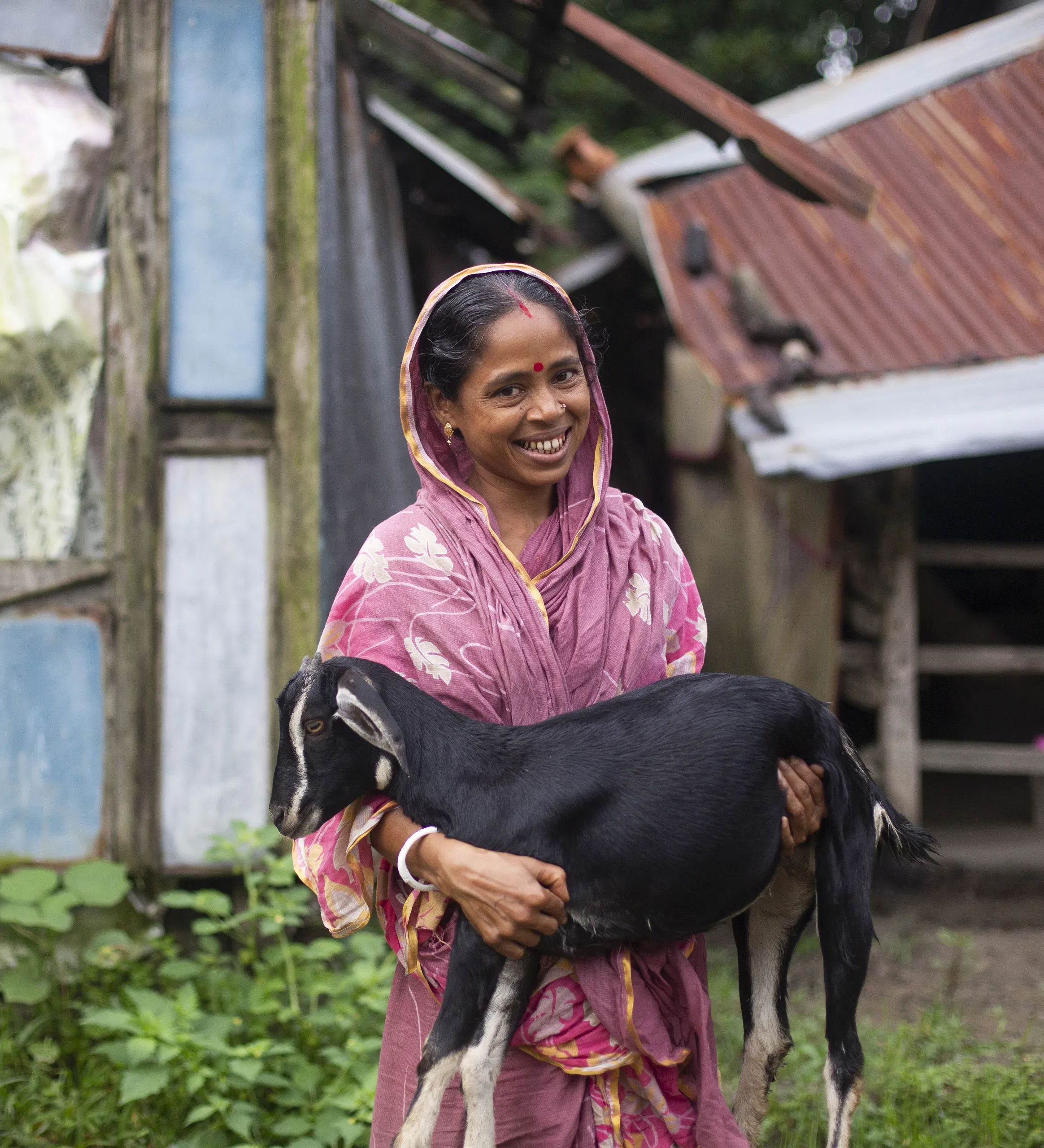 Woman in traditional attire holding a goat, standing in front of an old rustic house, smiling warmly.