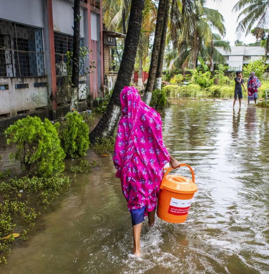 Woman in pink dress walking through flooded area carrying an orange bucket, surrounded by palm trees and greenery.