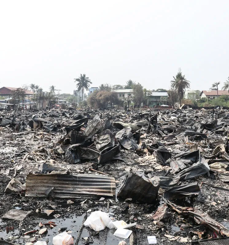 Charred debris and ruins after a fire in a residential area, with damaged corrugated sheets and palm trees in the background.