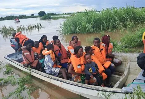 People in orange life vests sitting in a boat navigating floodwaters, surrounded by greenery and cloudy skies.