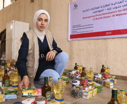 Woman organizing emergency food supplies in Gaza, surrounded by essential items under a relief banner.