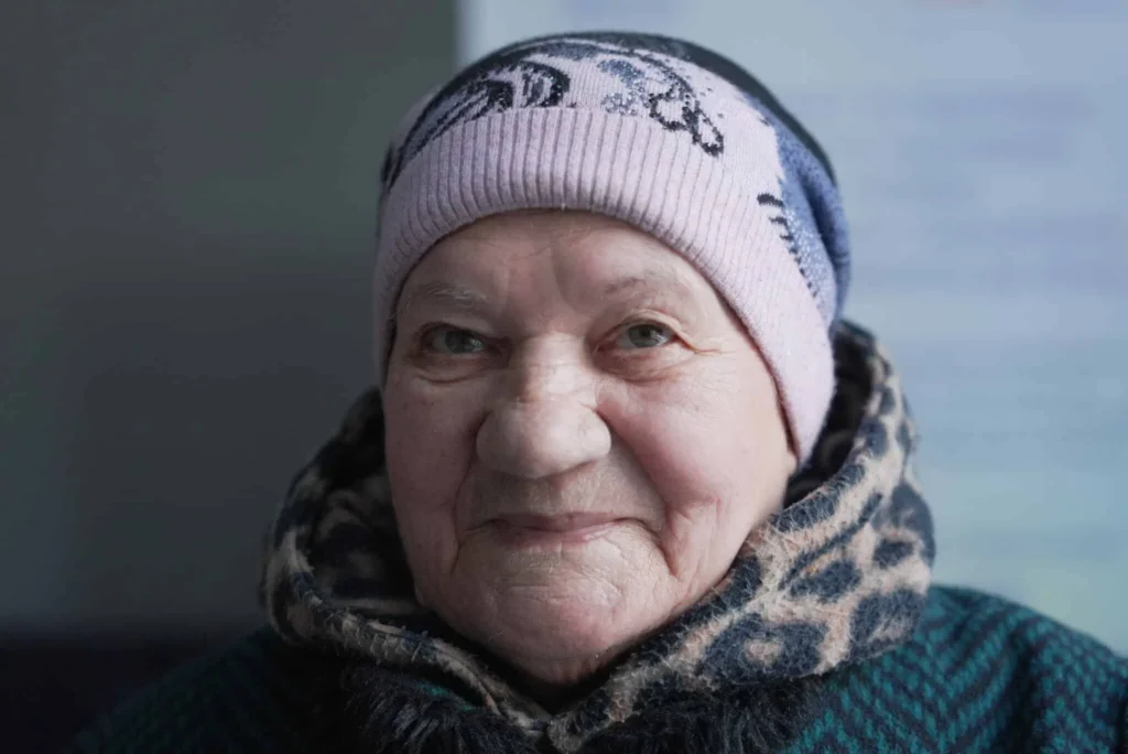 Elderly woman with a warm smile, wearing a knit hat and scarf, conveying warmth and kindness.
