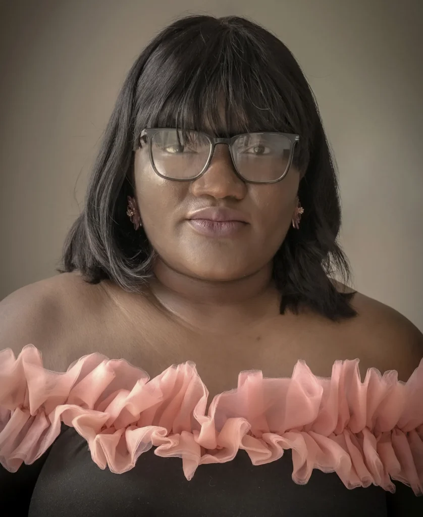 Woman in glasses wearing an off-shoulder black dress with ruffled pink trim, looking confidently at the camera.