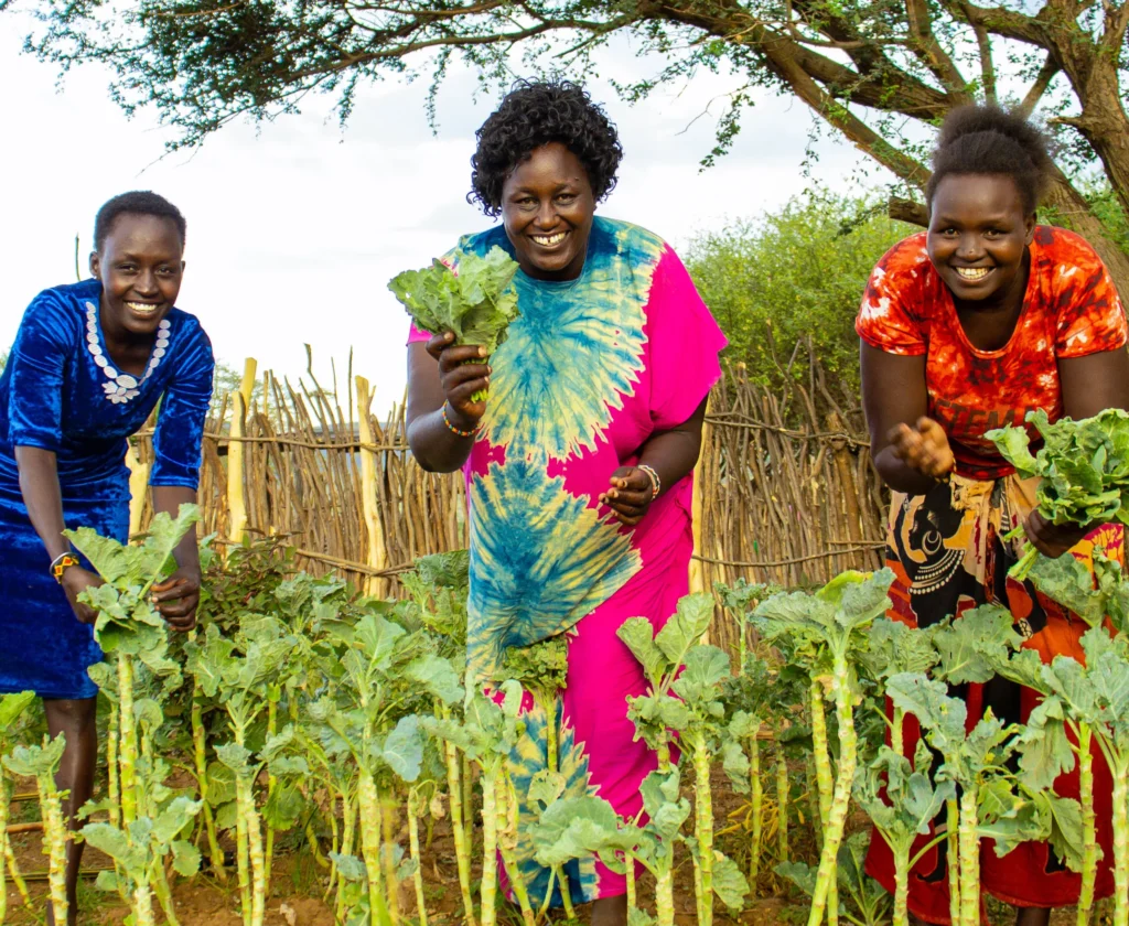 Three women smiling and harvesting leafy greens in a vibrant community garden.