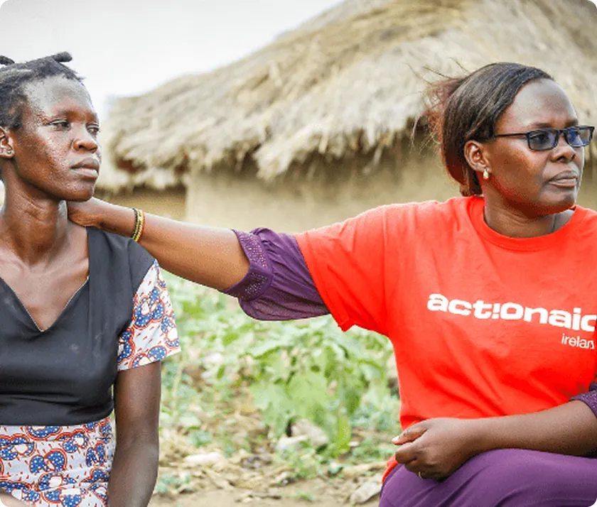Two women sit together outside a hut, one offering comfort with a hand on the other's shoulder.