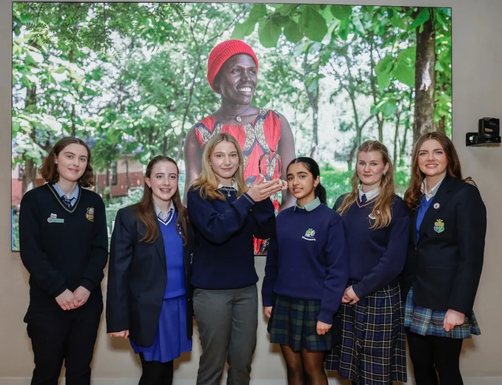 Group of students in school uniforms stand in front of a vibrant nature-themed display screen.
