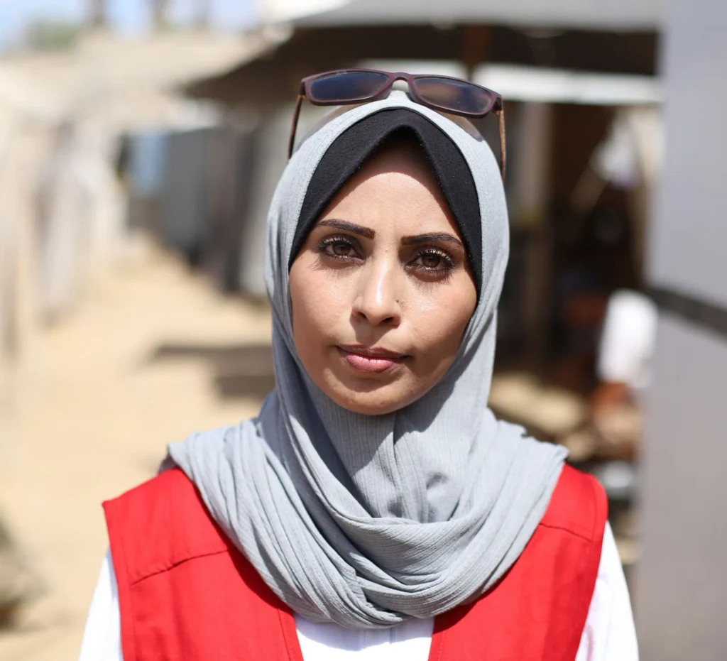 Young woman in gray hijab and red vest outdoors, looking directly at the camera, with blurred background.