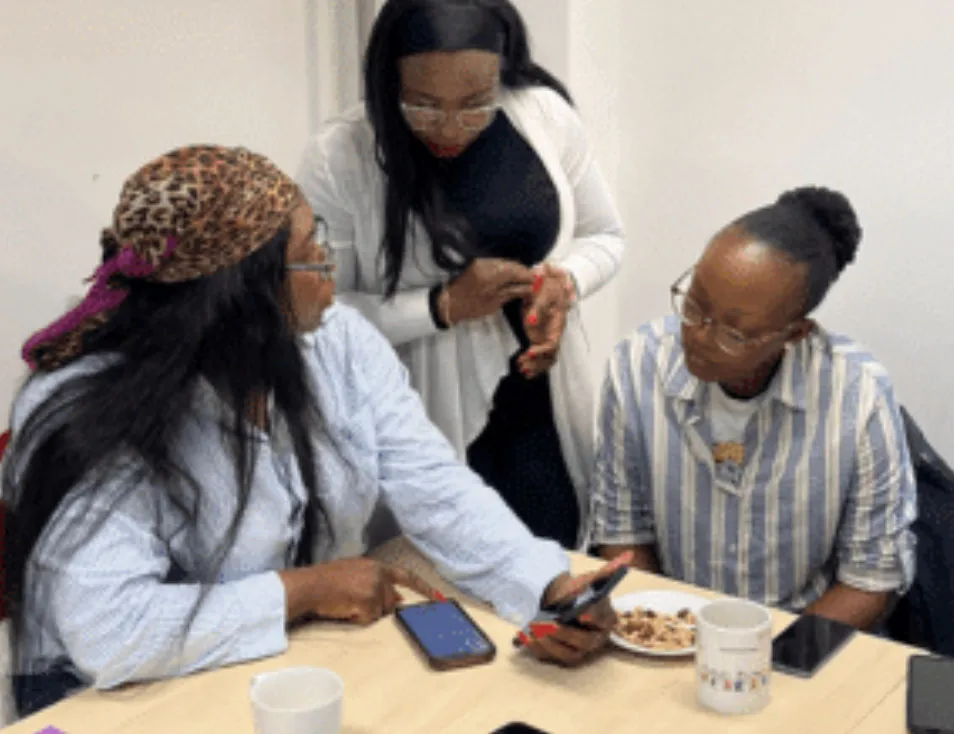 Three women collaborating in a meeting, with one pointing at a phone on a table with snacks and mugs.
