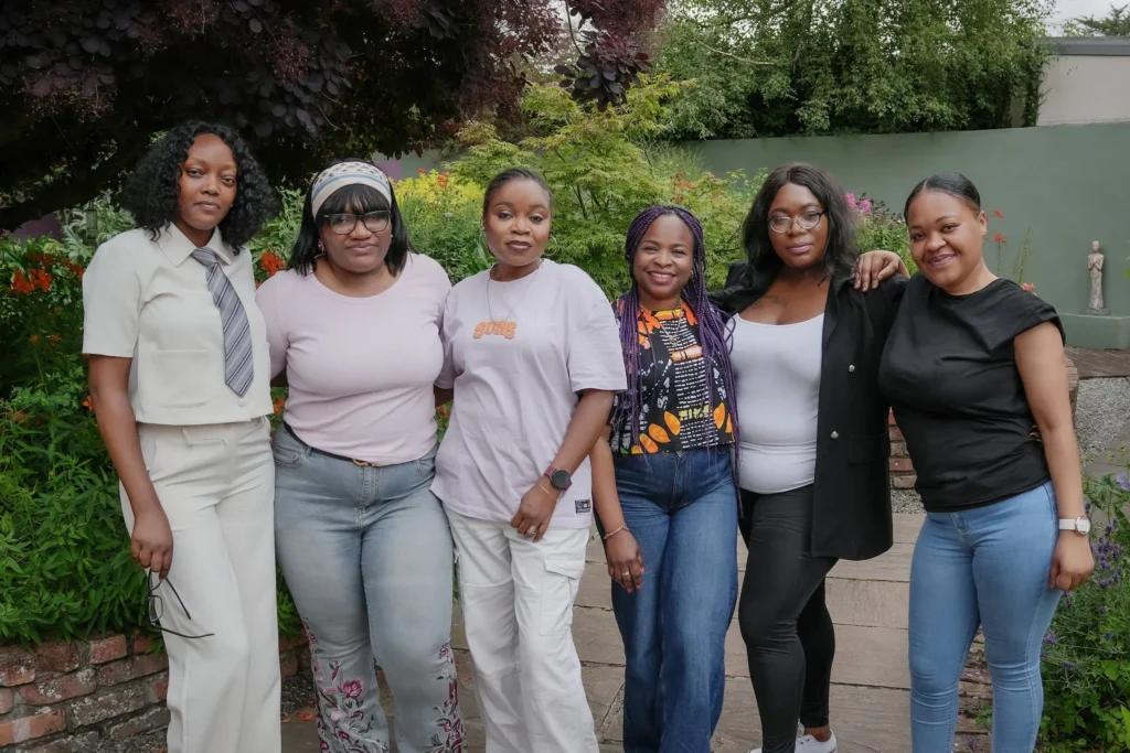 Group of six women smiling outdoors, standing in garden with colorful flowers and greenery.