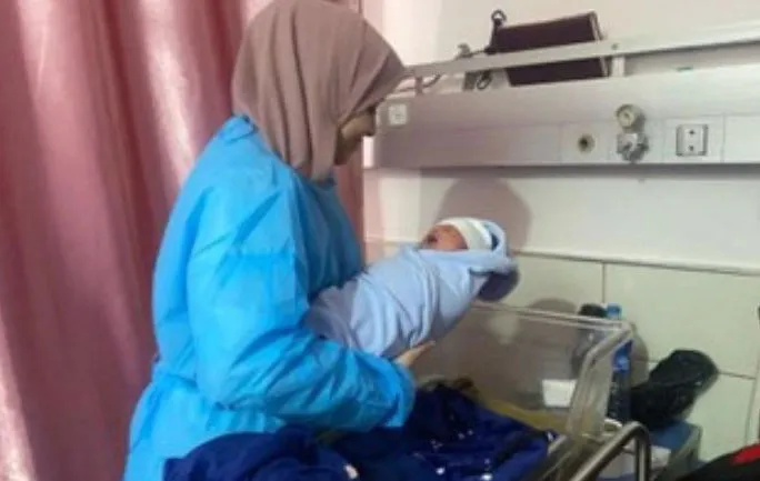 Healthcare worker in blue attire gently holding a newborn wrapped in a blanket at the hospital, next to baby crib.