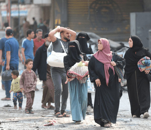 People walking through a street, carrying bags, amid rubble and damage, suggesting a challenging environment or crisis.