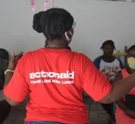 A person in a red ActionAid shirt interacts with a group during a community event indoors.