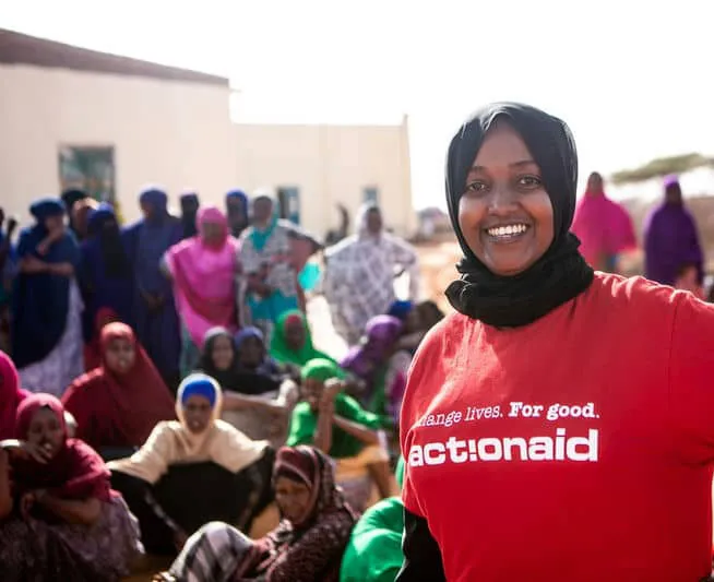 Smiling woman in ActionAid shirt stands in front of a group of people outdoors, promoting empowerment and community support.