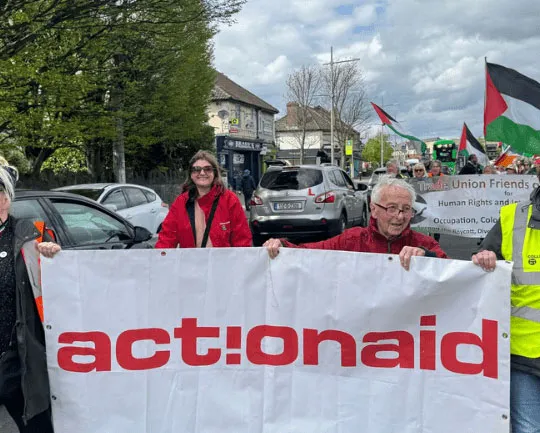 Protesters hold ActionAid banner during a street demonstration, surrounded by flags and signs on a cloudy day.