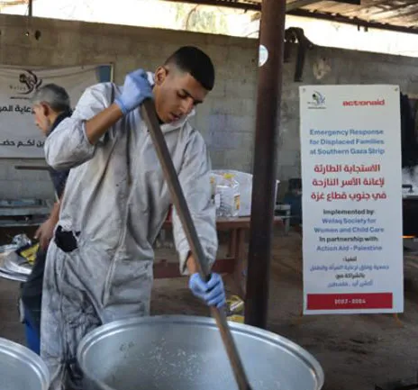 Man stirring large pot during emergency response for displaced families in Southern Gaza Strip, with ActionAid sign in background.