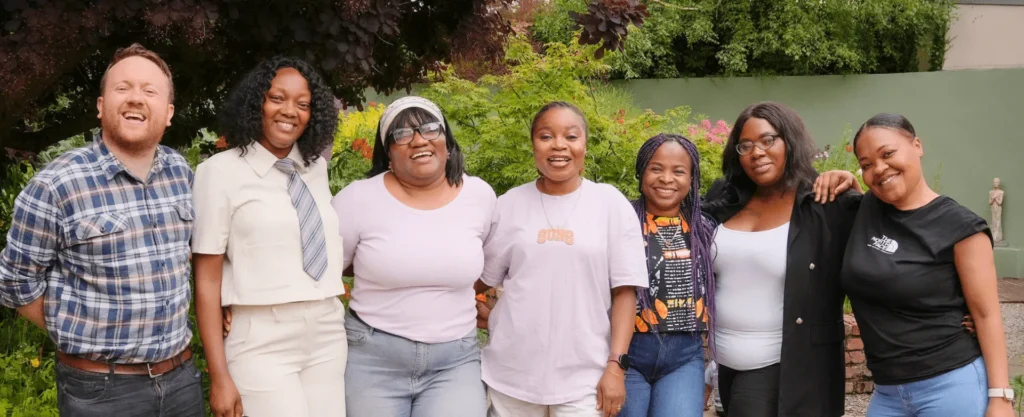 A diverse group of seven people smiling outdoors with greenery in the background.
