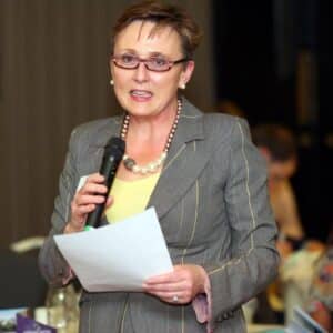 Woman in a gray suit speaking into a microphone at an event, holding a paper, with an audience in the background.