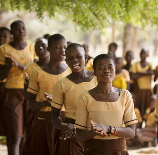 Group of students clapping and singing outdoors in uniform, under trees, engaging in a musical activity.