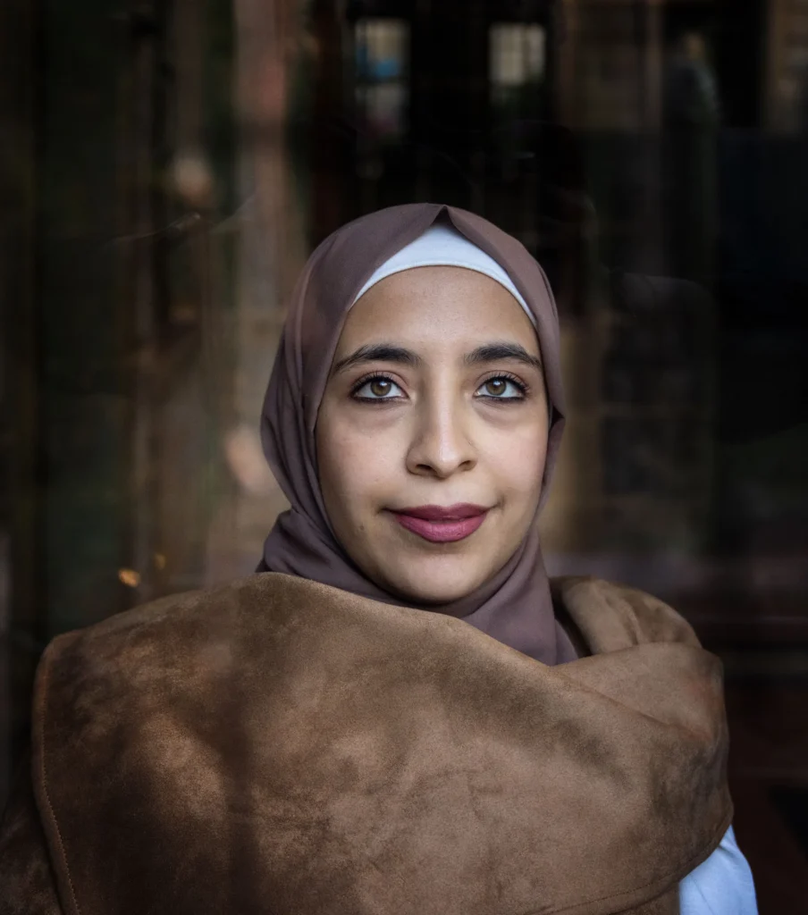 Portrait of a woman in a brown hijab, with a soft smile and a blurred background.