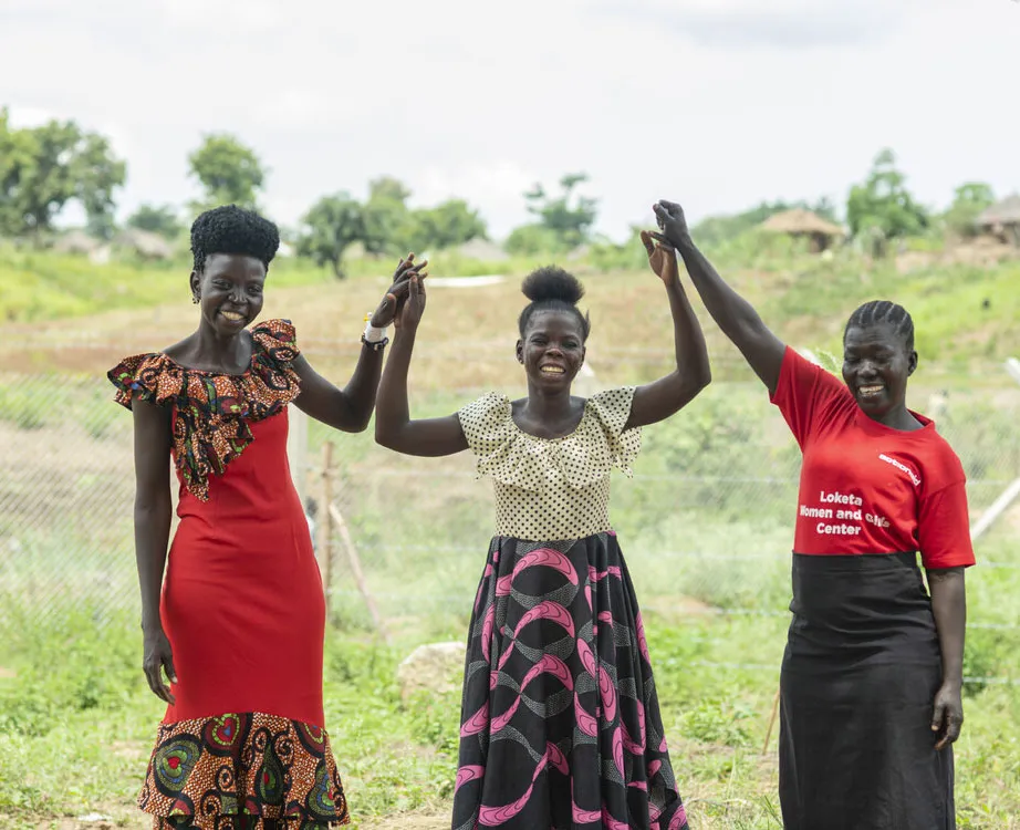 Three women holding hands and smiling outdoors, promoting community empowerment and unity.