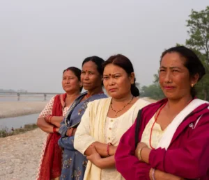 Four women stand united on a riverbank, looking ahead with determination under a cloudy sky.