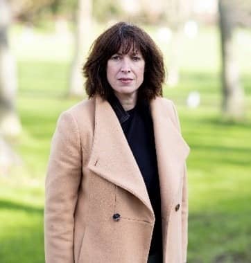 Woman in a tan coat standing in a sunny park with blurred trees in the background.