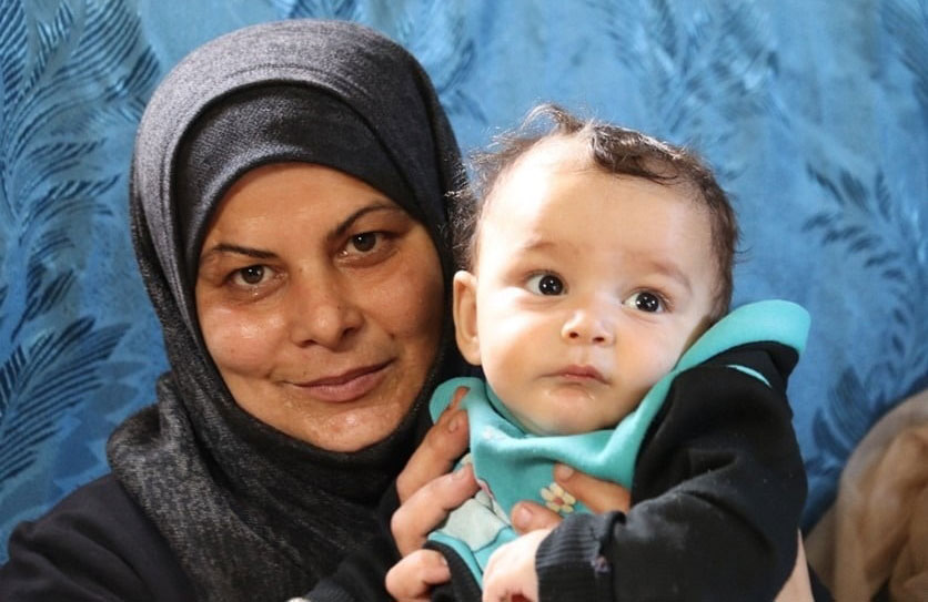 Woman in hijab smiling, holding an infant with a turquoise bib against a blue background.