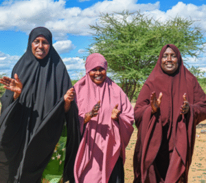 Three women in colorful hijabs smile and clap in a sunny outdoor setting with trees and blue sky in the background.