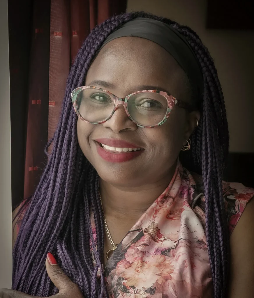 Portrait of a woman with purple braids, floral top, and glasses, smiling by a curtain in natural light.