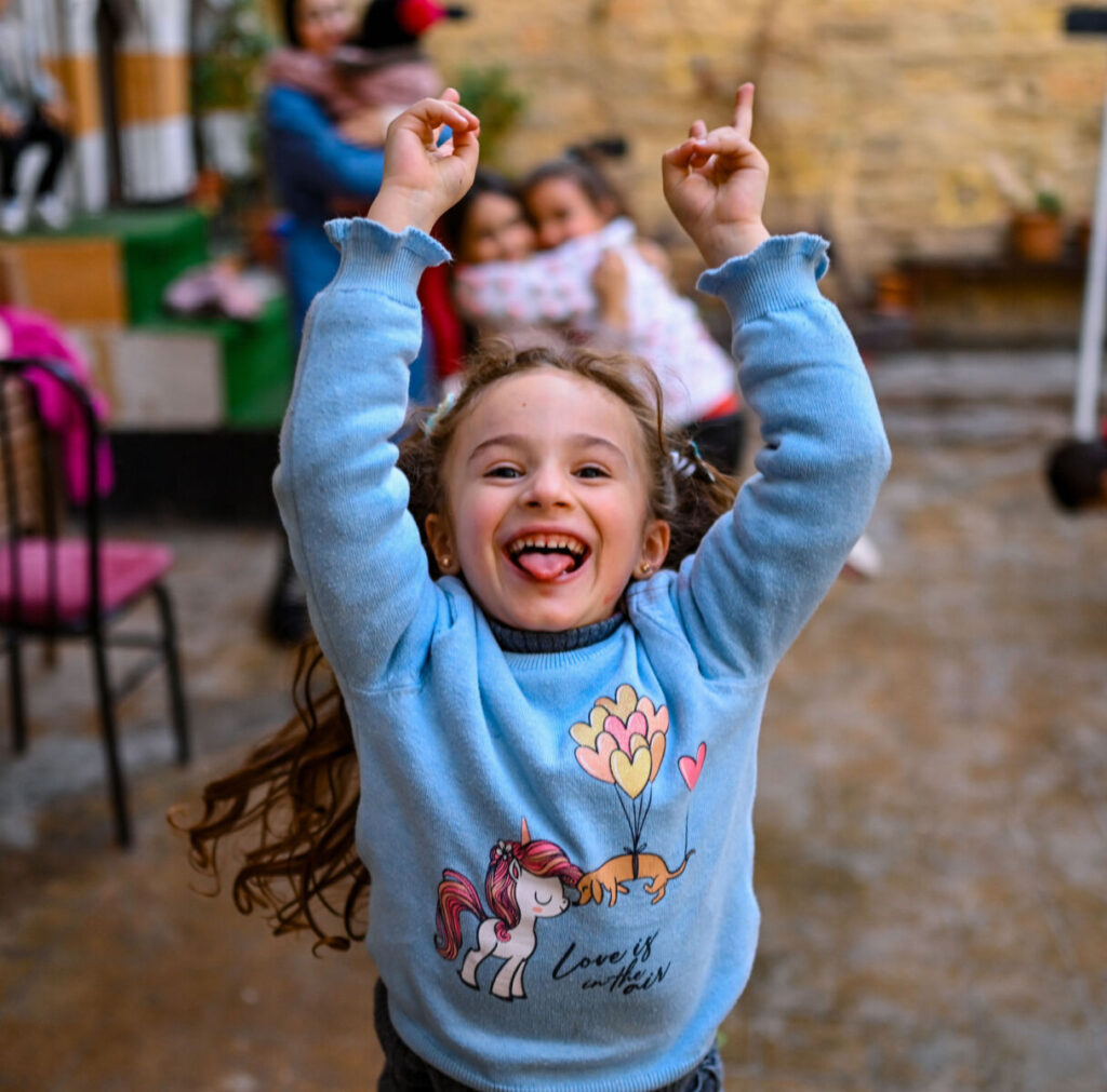 Joyful child in blue sweater with unicorn, smiling and raising arms outside, embodying happiness and playfulness.