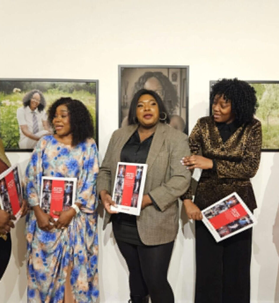 Group of women holding books at an art exhibit, standing by framed portraits on the wall.