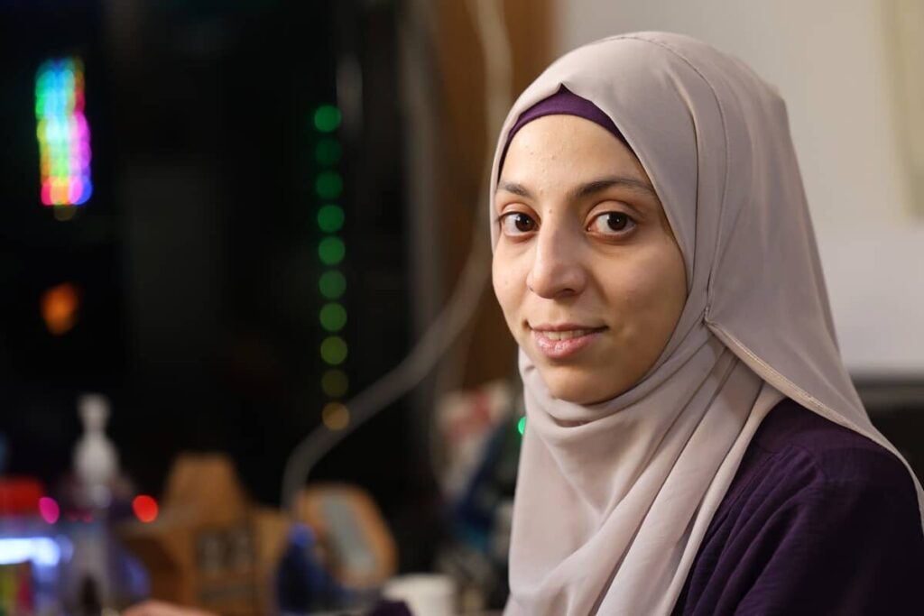 Woman in beige hijab and purple shirt smiling softly indoors.