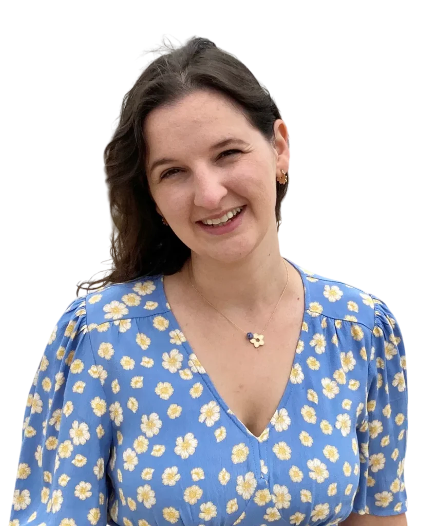 Smiling woman in a blue floral dress with a daisy necklace, standing against a plain background.