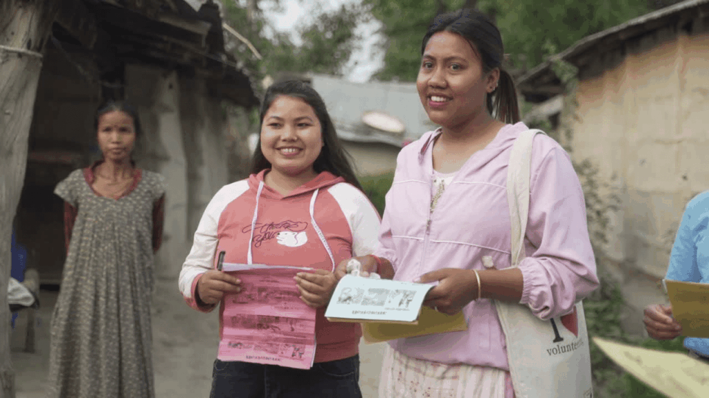 Two women smiling while distributing flyers in a village setting, engaged in community outreach.