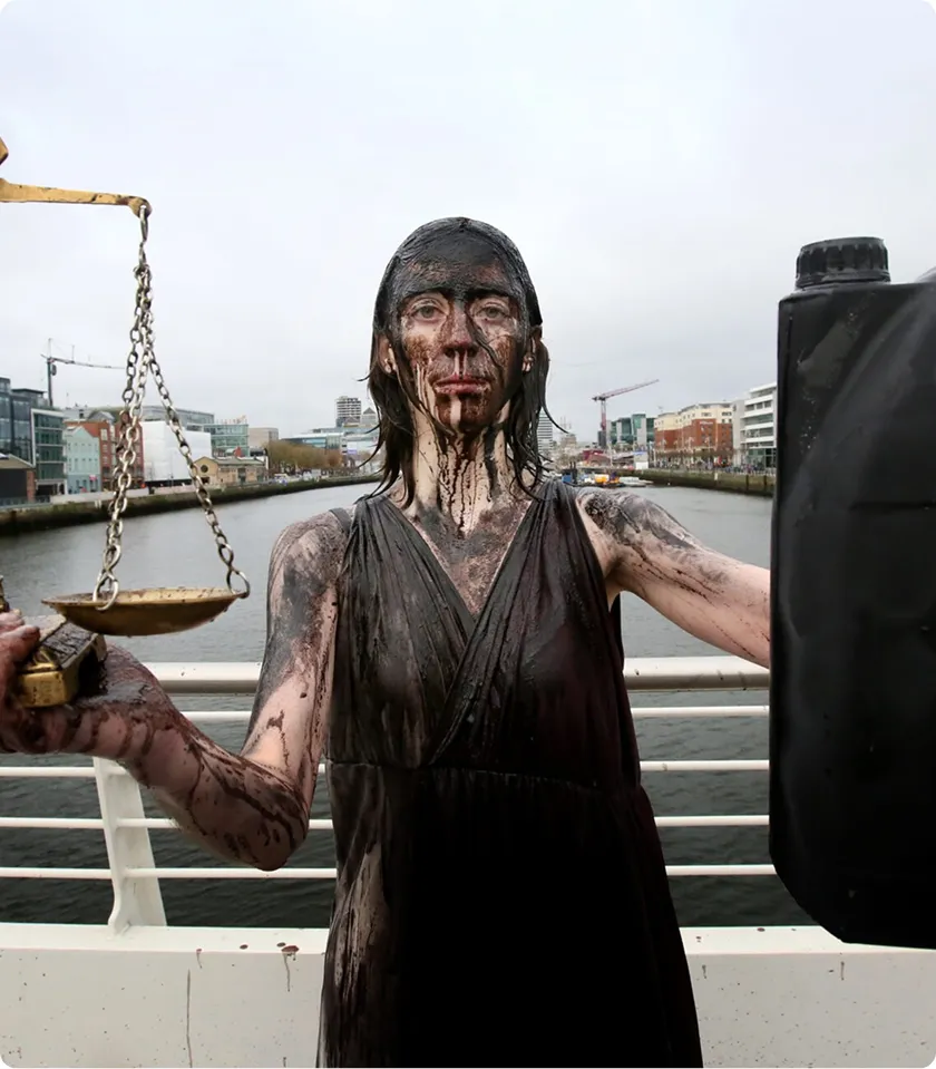 Protester covered in oil-like substance holding scales and canister, symbolizing environmental activism by the river.